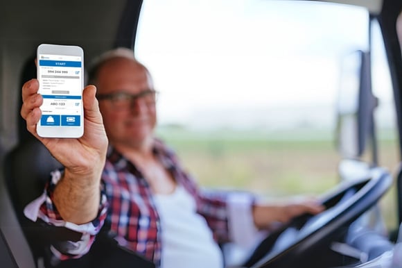 An older male truck driver sitting inside the cabin, holding up a smartphone towards the camera. The phone screen displays the mScales user interface.