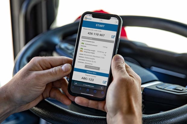 Smartphone in the driver’s hand displaying the mScales start screen, with a truck steering wheel and dashboard in the background.