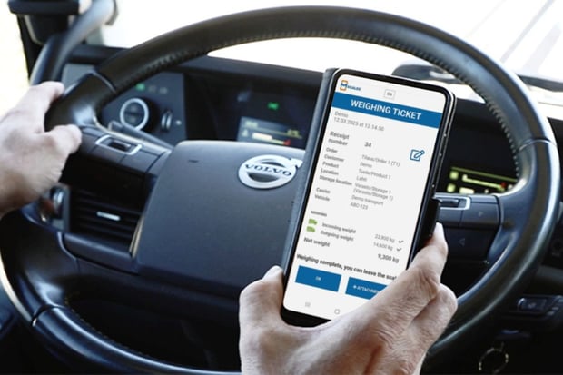 Smartphone in the driver’s hand displaying an mScales weighing ticket, with a truck steering wheel and dashboard in the background.