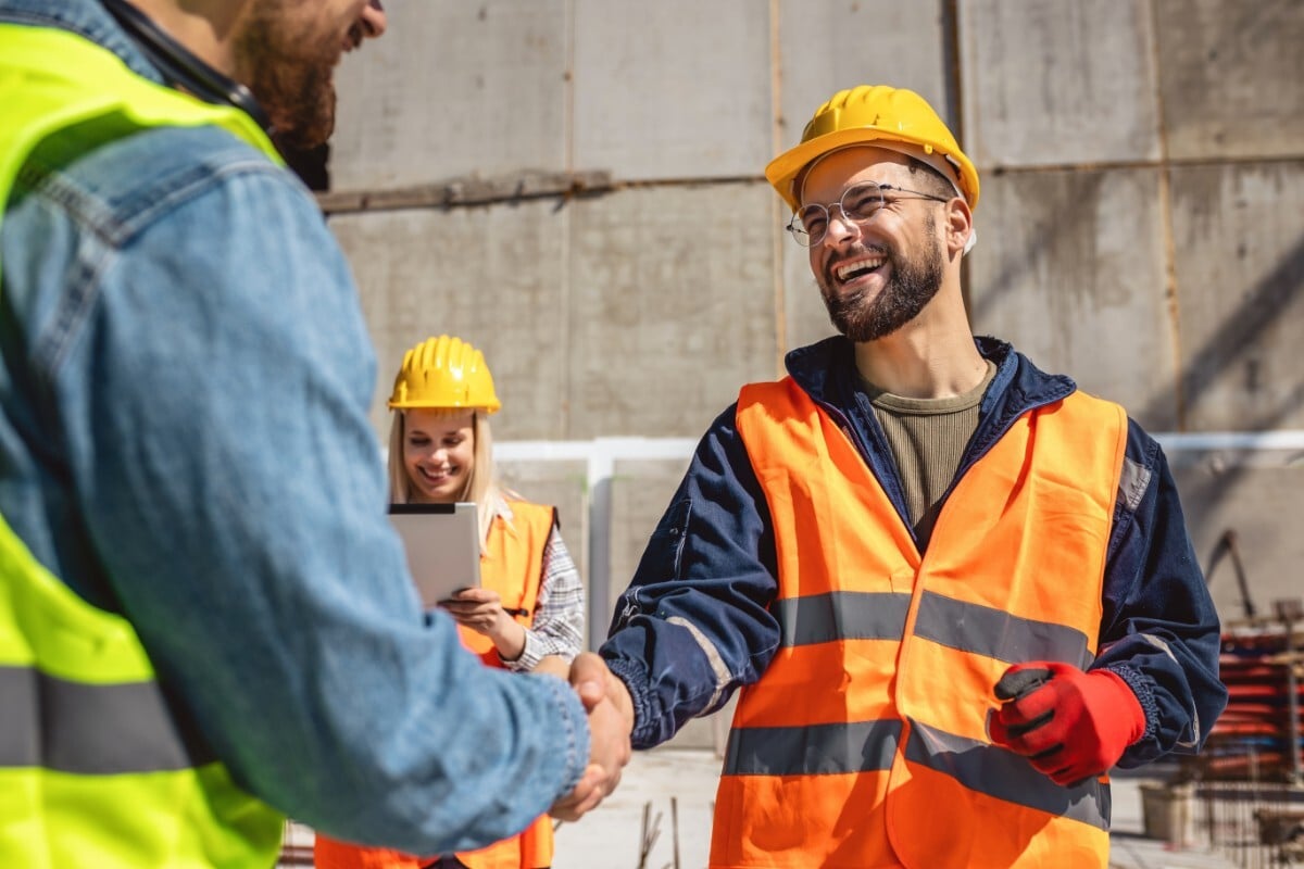 Two workers wearing safety vests and helmets shaking hands and smiling outdoors in bright sunlight