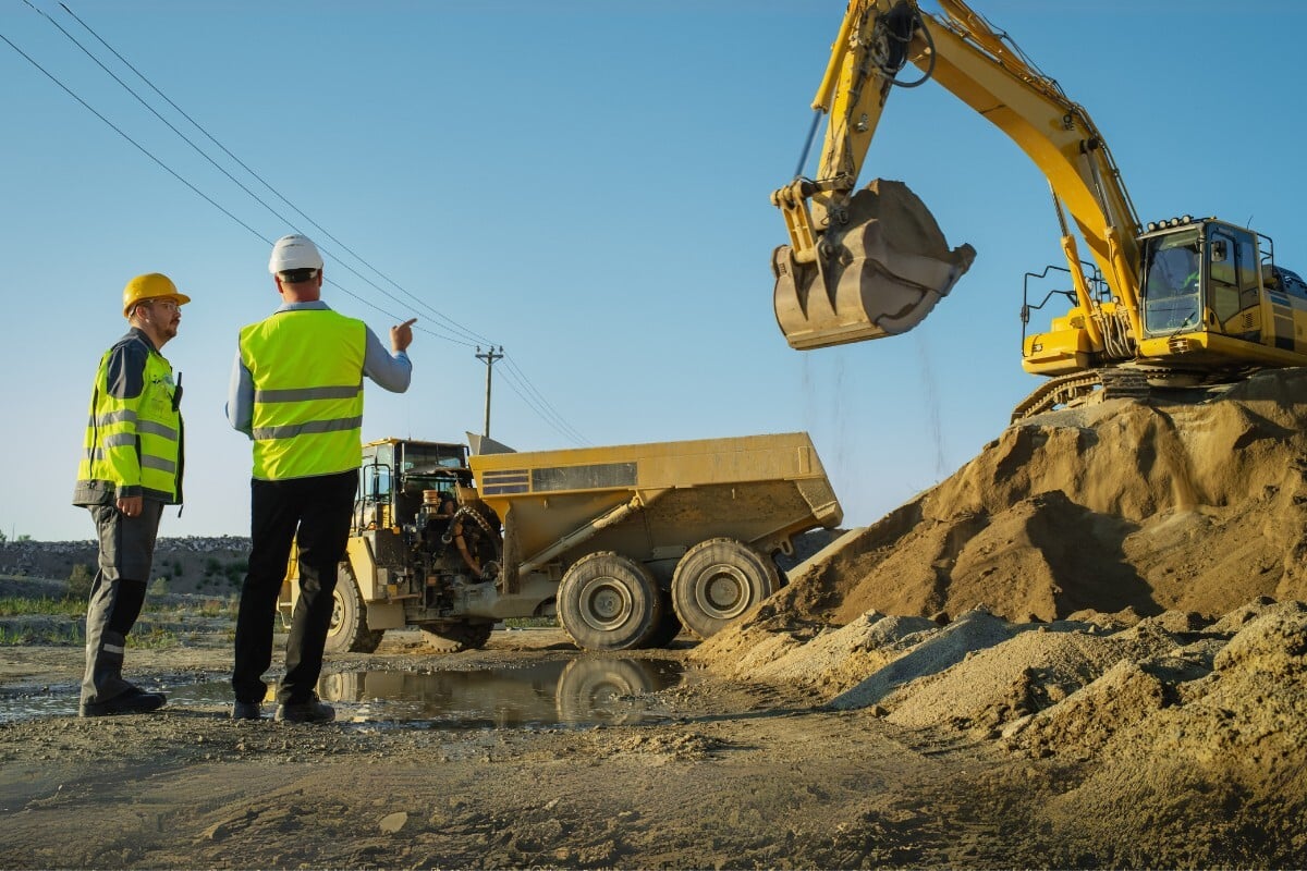 Two workers in high-visibility clothing talking near a wheel loader and dump truck beside a sand pile on a sunny day with a blue sky