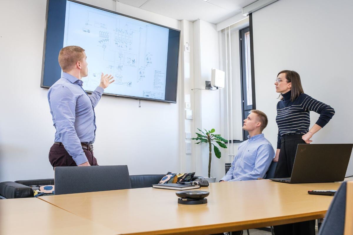 Professional meeting in a modern office where a man presents a technical diagram on a large screen while two colleagues listen, with a laptop and plant on the conference table indicating a collaborative work setting.