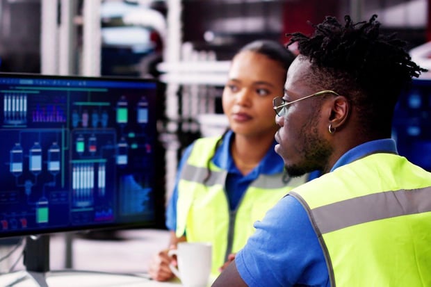 Two individuals in safety vests working in a technical environment, focused on a computer screen displaying data, suggesting collaboration in monitoring or analyzing operations.
