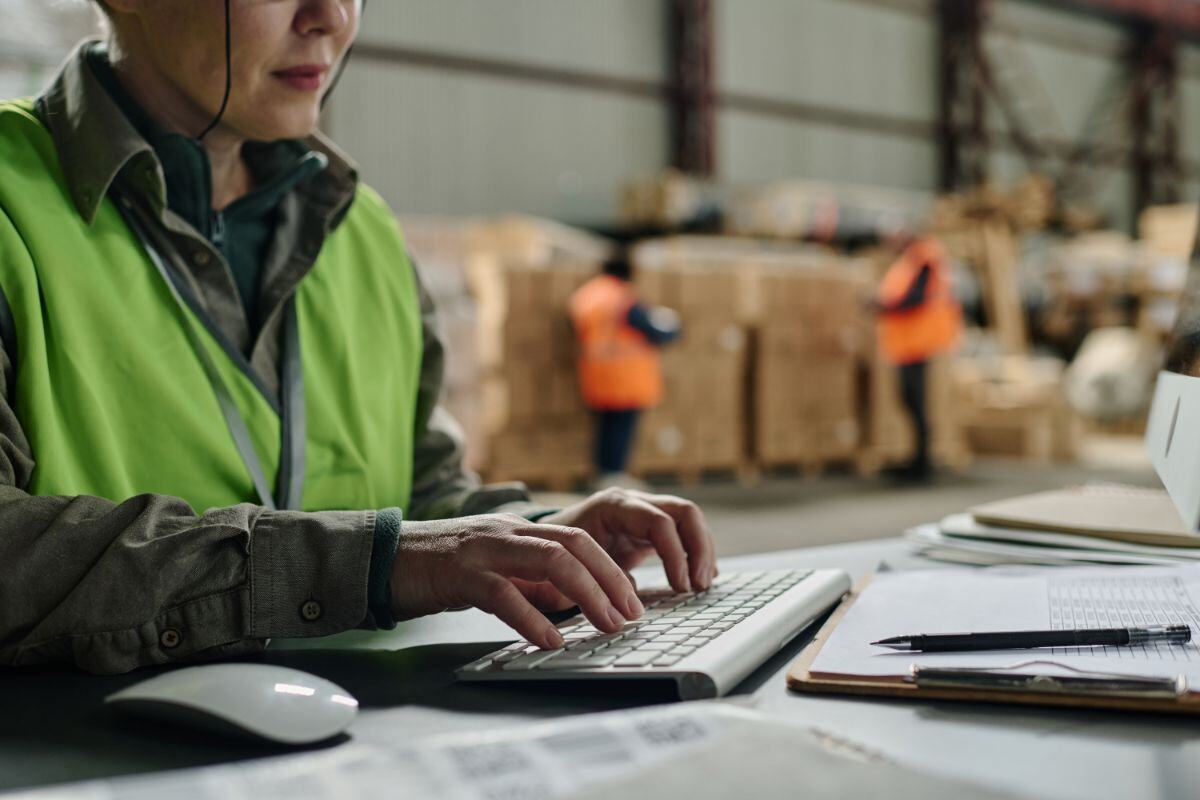 Person wearing a bright green safety vest typing at a keyboard in a warehouse, with other workers and stacks of boxes in the background, indicating a logistics or distribution environment.