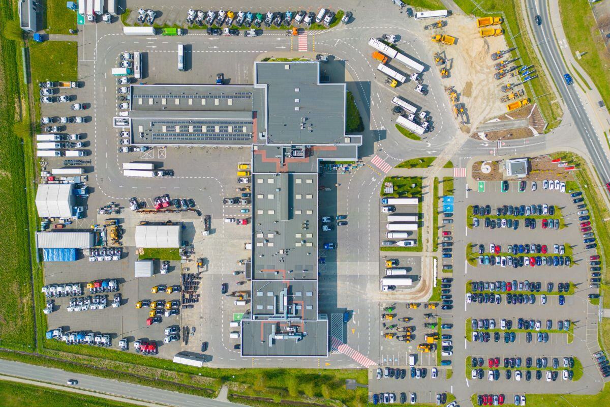 Aerial view of a large logistics or distribution center with a flat-roofed central building, surrounding parking areas for cars and trucks, access roads, loading zones, and construction vehicles, set within a structured layout and green surroundings.