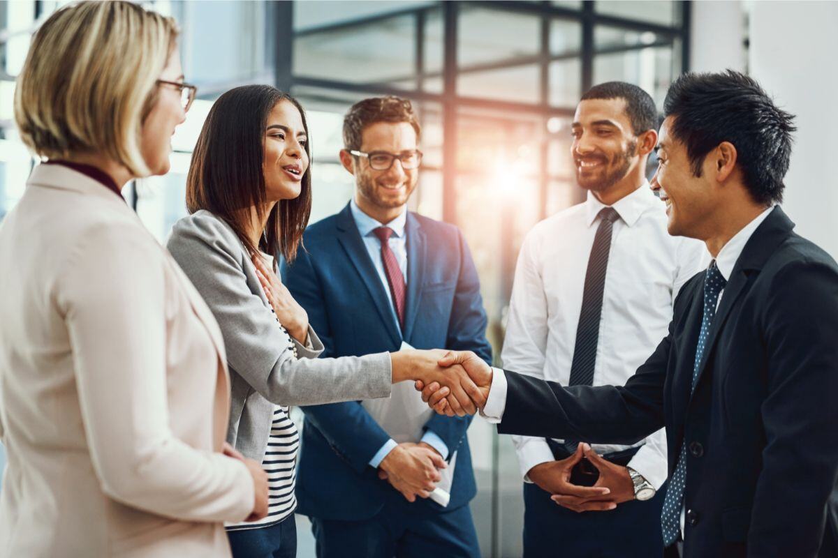 Group of professionals in a modern corporate setting, with a woman in a gray blazer shaking hands with a man in a suit as others look on and smile, conveying a positive and collaborative atmosphere.