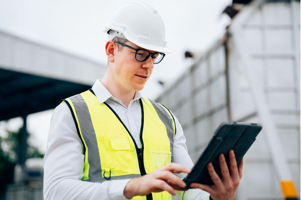 Man wearing a white hard hat and yellow safety vest using a tablet at a construction site, with a partially built structure in the background.