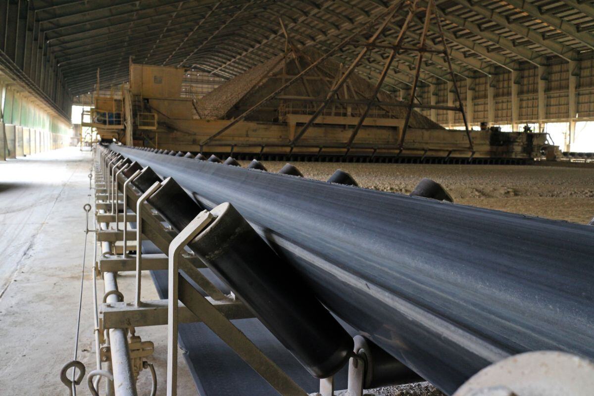Industrial facility with a long conveyor belt system supported by metal structures, leading toward large machinery and a pile of aggregate or bulk materials in an enclosed processing or storage area.