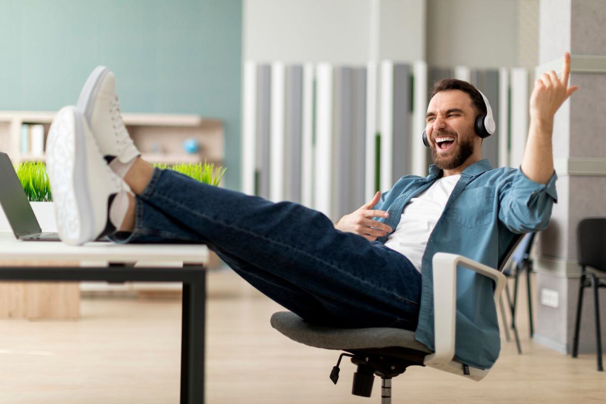 Man in a modern office wearing headphones, laughing and gesturing with one hand while relaxing with his feet on a table, in a casual setting with greenery and contemporary furniture