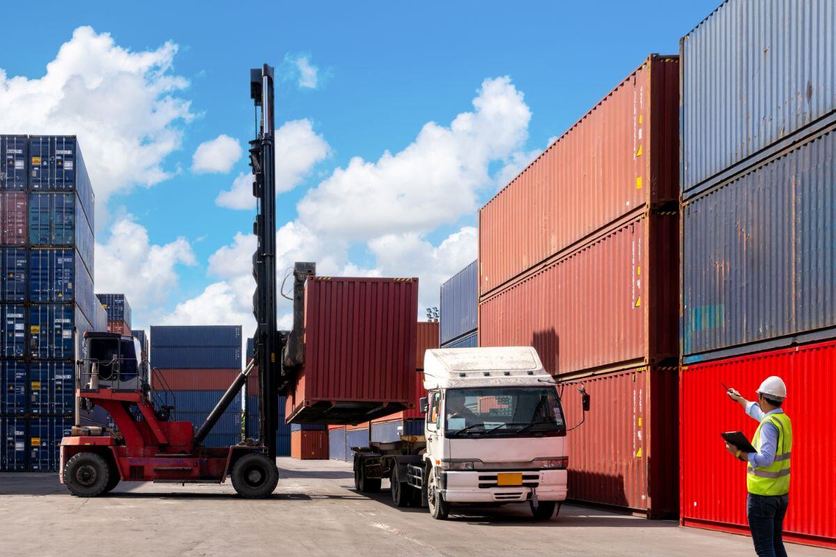 Large red forklift lifting a shipping container to load onto a white truck in a busy shipping yard, with stacked containers in the background and a worker in a hard hat directing the operation under a clear blue sky.