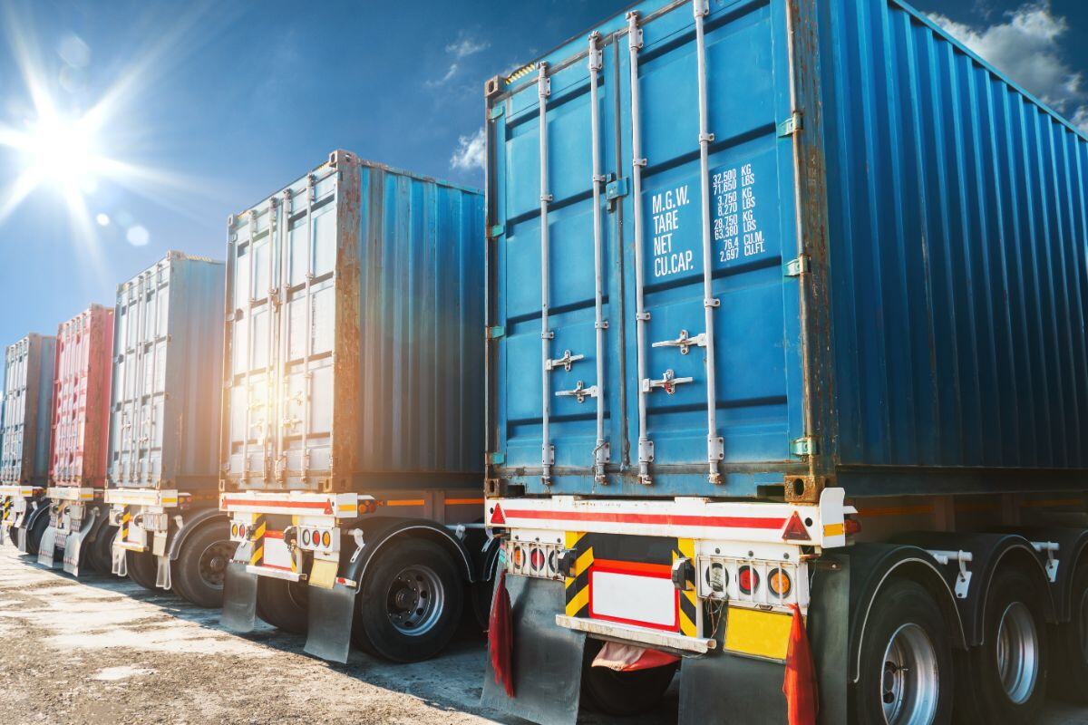 Row of large blue shipping containers stacked on trucks under a bright sunny sky, with visible labels and measurements, illustrating industrial logistics and freight transportation.