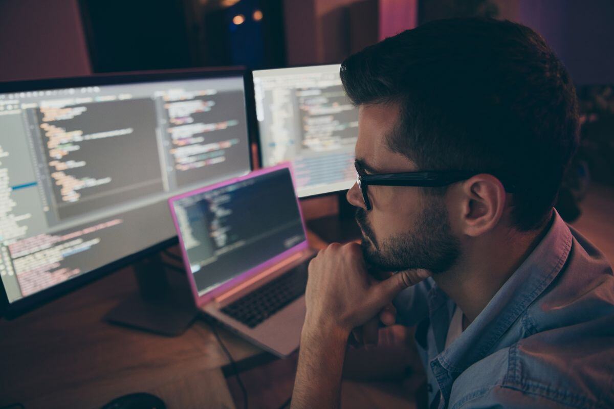 Person sitting at a desk in a dimly lit workspace, surrounded by multiple screens displaying code, with one hand on their chin, suggesting deep focus in a programming or software development environment.