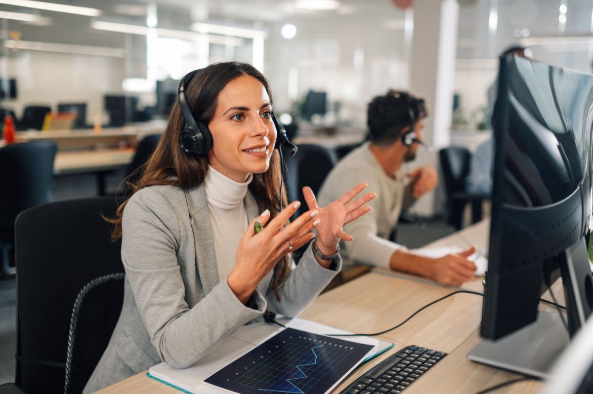 Woman wearing a headset in a professional setting, speaking while seated at a desk with a computer and a tablet displaying a graph, with a colleague visible in the background in a focused office environment.