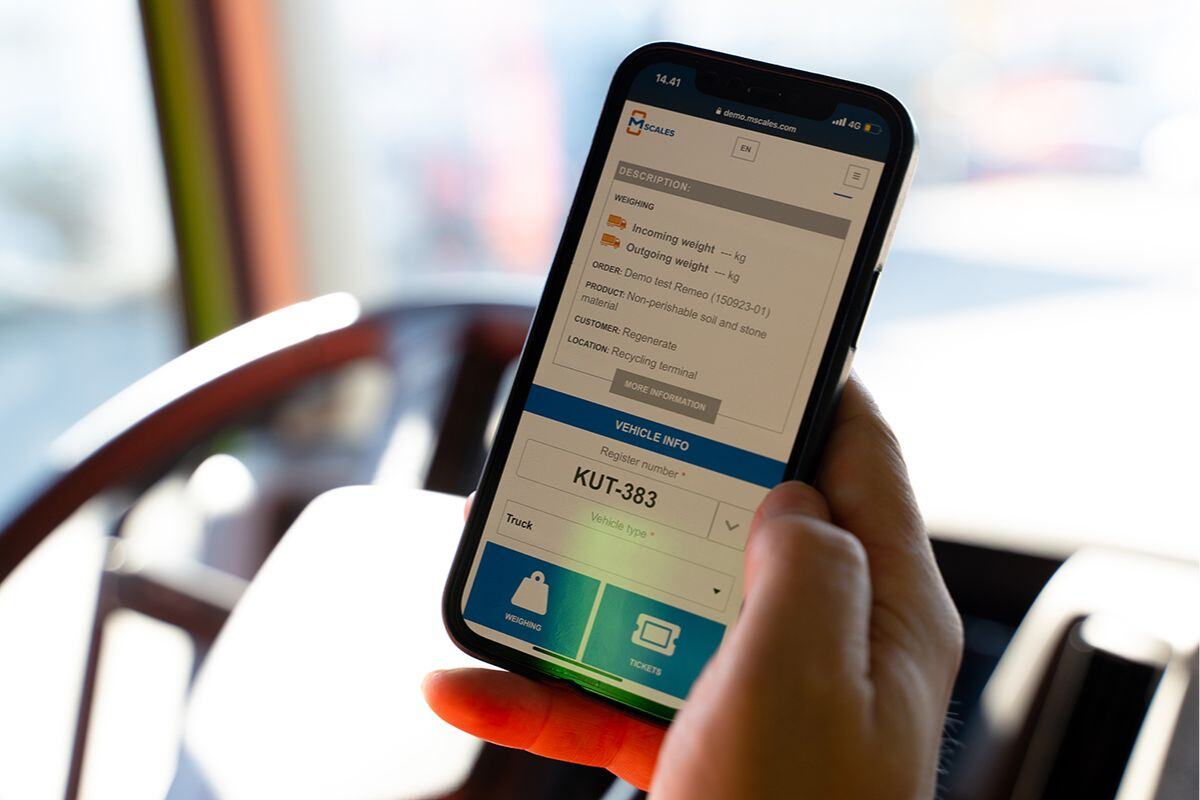 Close-up of a hand holding a smartphone displaying the mScales user interface for vehicle weighing, with fields for weight and product data visible, inside a truck.
