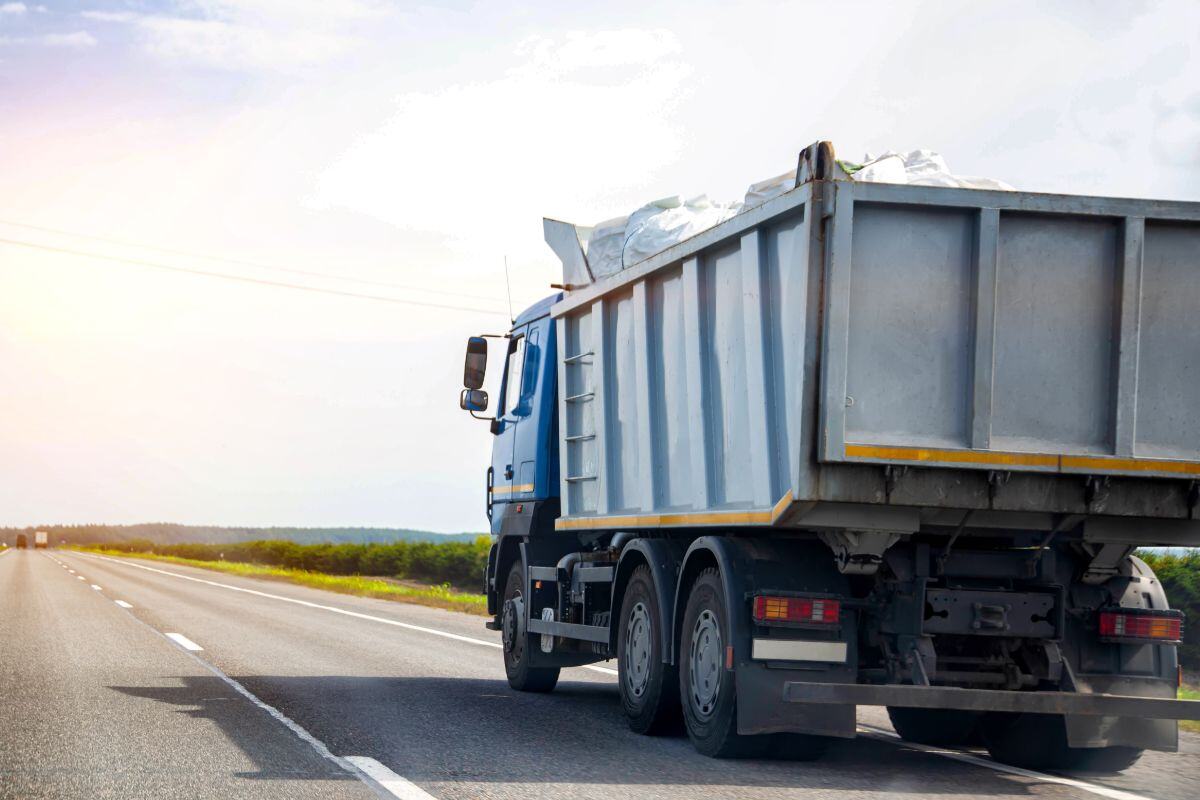 Blue dump truck traveling on a highway with a loaded bed, set against a partly cloudy sky and green fields