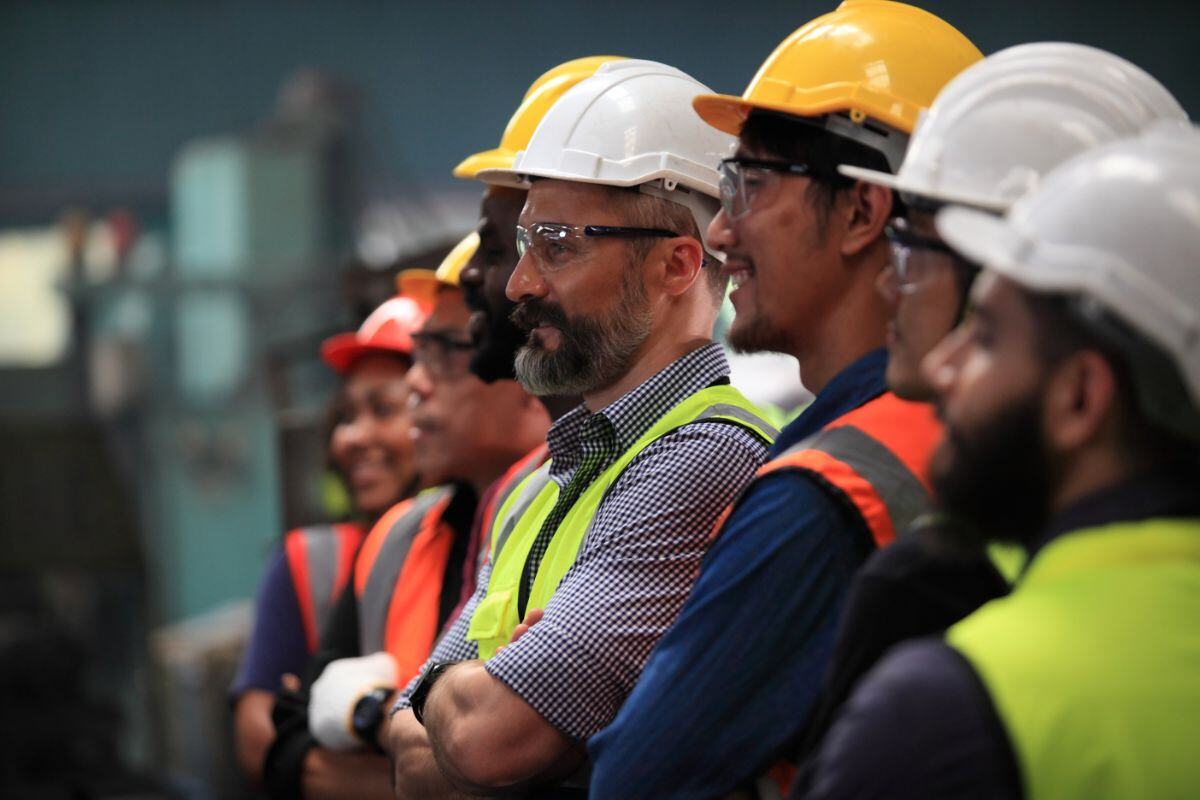 Group of workers in safety helmets and vests in a manufacturing or construction setting, appearing engaged and attentive during a training or informational session, with expressions ranging from focused to smiling.