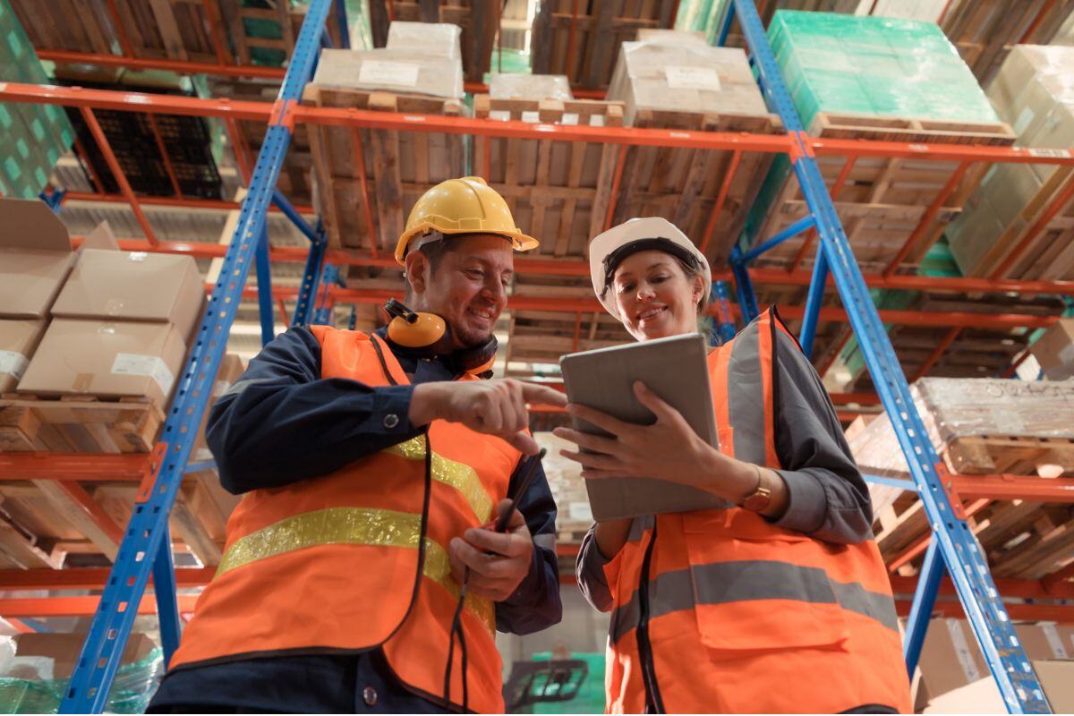 Two warehouse workers wearing high-visibility vests and hard hats discussing a task while looking at a tablet, with shelves of boxes in the background in a warehouse environment.