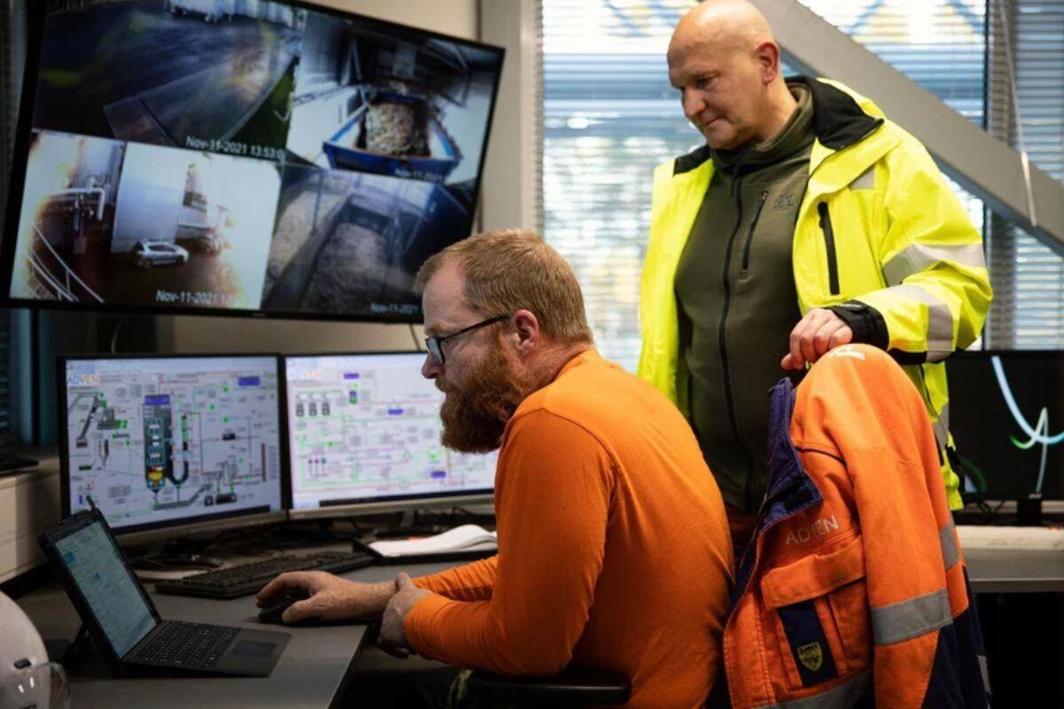 Two individuals in a control room, one seated in an orange shirt working on a laptop with operational data, and the other standing in a yellow safety jacket observing screens showing surveillance footage and system controls in an industrial facility.