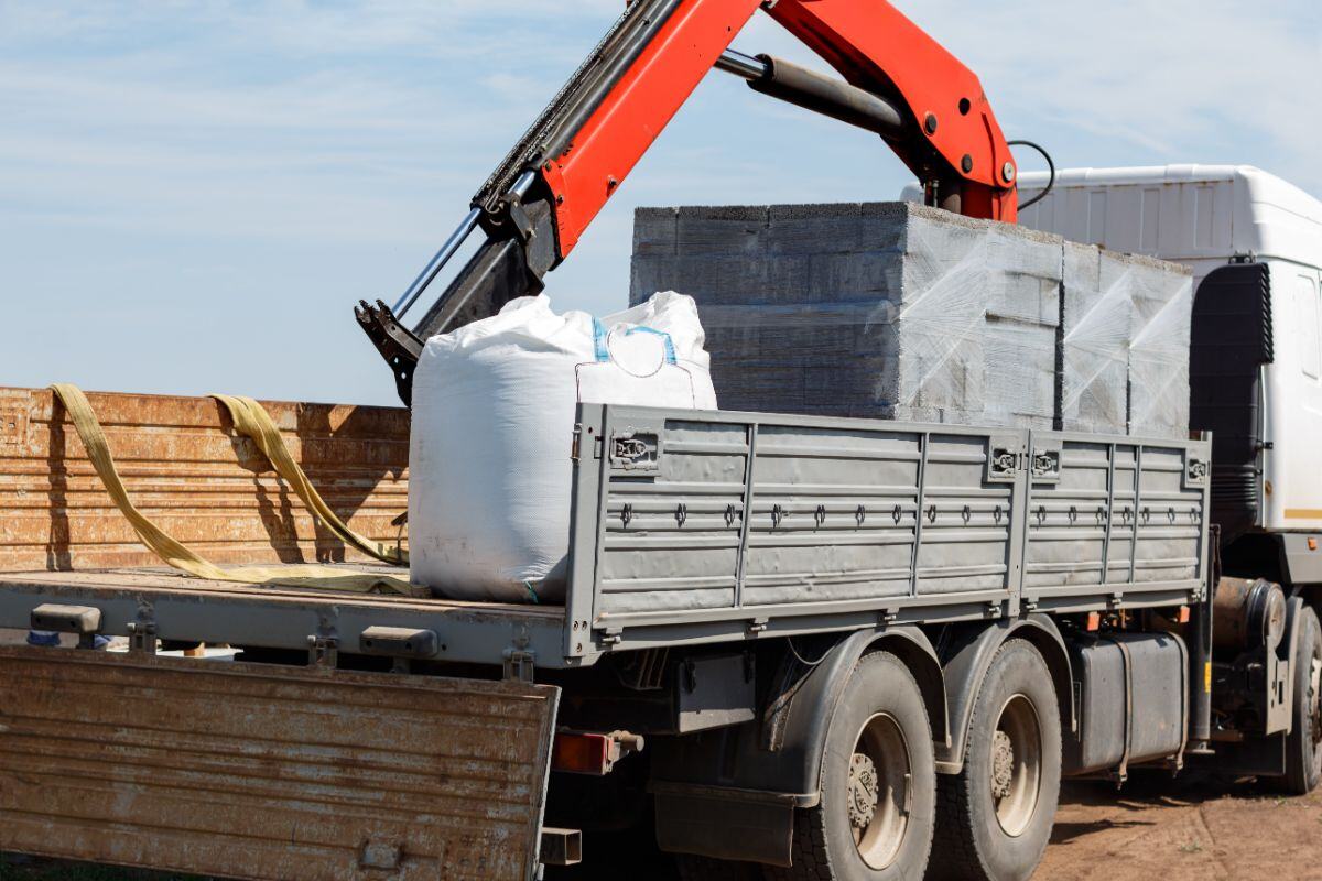 Truck with a flatbed loaded with gray construction blocks and a large white bag, with a crane positioned above, set outdoors on a construction site under a clear blue sky.
