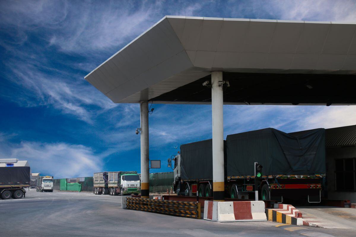 Truck terminal or checkpoint with several parked trucks under a large canopy, traffic control signals, marked lanes, and barriers, set against a blue sky with clouds.