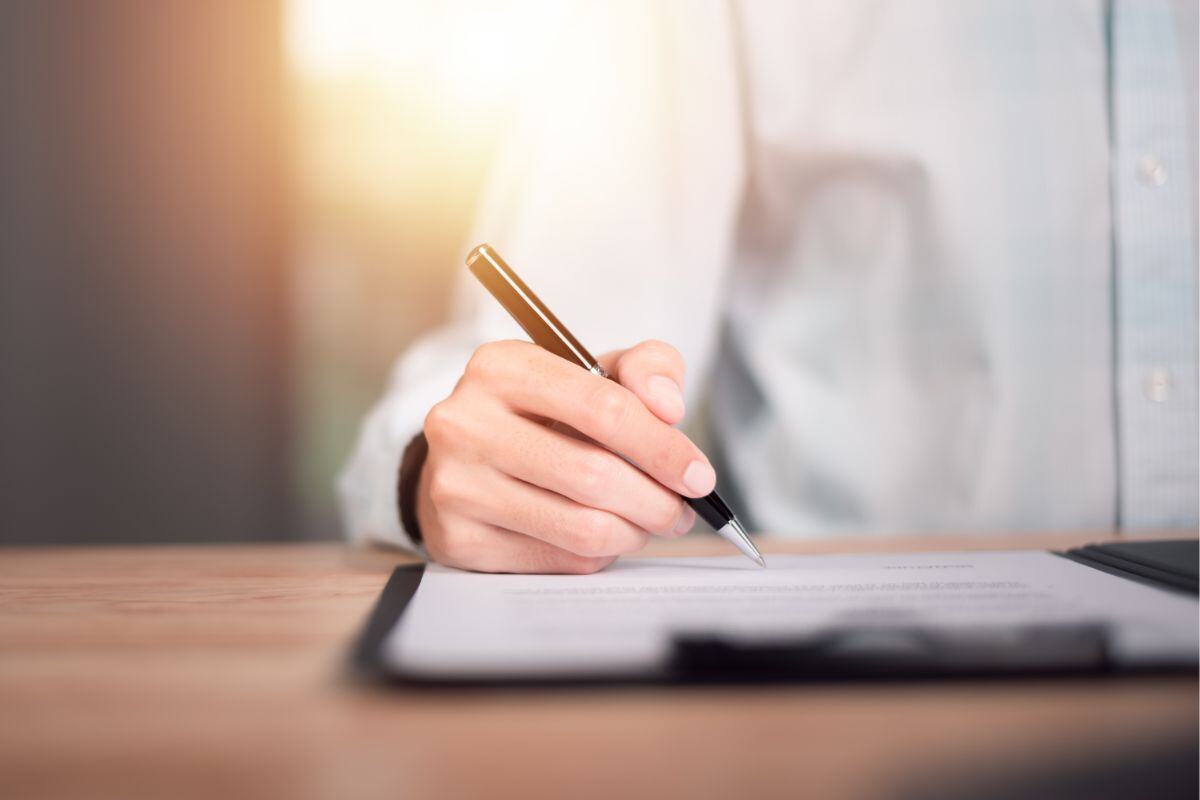Close-up of a hand holding a pen over a document on a clipboard, with warm light in the background suggesting a focused work or study environment